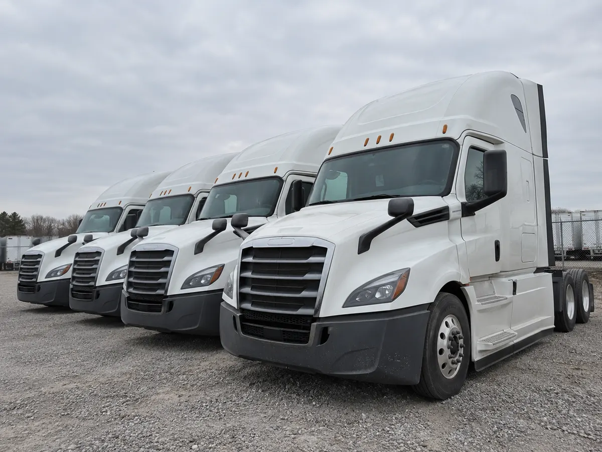 Four white semi tractors parked side by side in a gravel yard, their factory headlights facing the camera